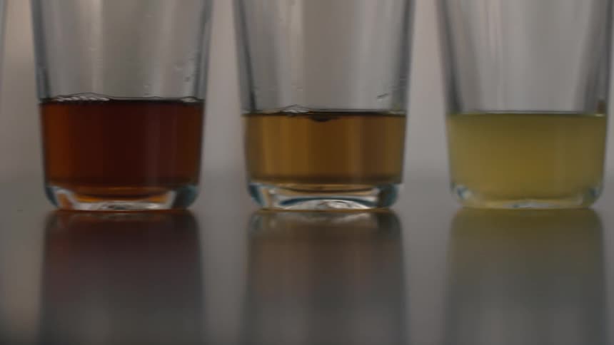 A panning shot reveals a line of clear glass tumblers on a reflective dark surface each containing a different liquidthe liquids range in color from a very dark brown or amber on the left to progressively lighter shades of brown yellowgold and pal