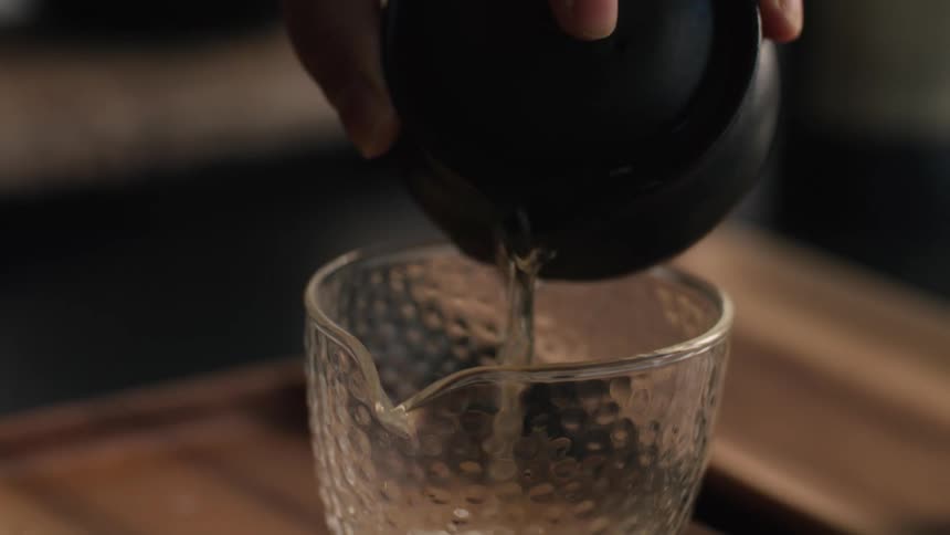 Male hands pouring tea from a black pot into a glass cup over a wooden tray on a table indoors