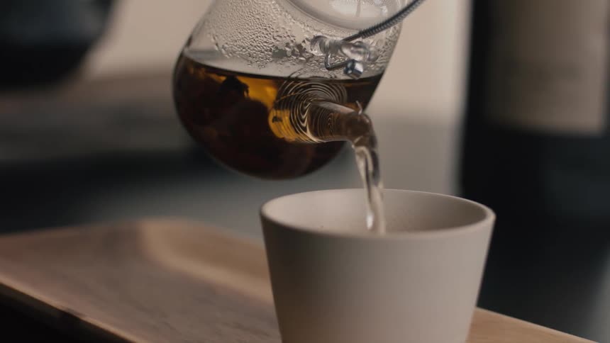 Hot tea being poured from glass teapot into white cup on wooden tray