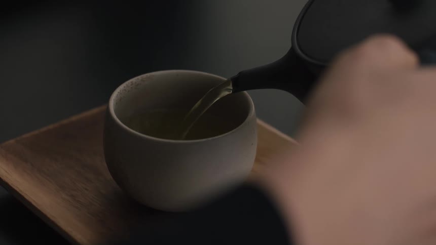 A womans hand pours tea from a dark teapot into a small matte gray ceramic cup sitting on a wooden tray