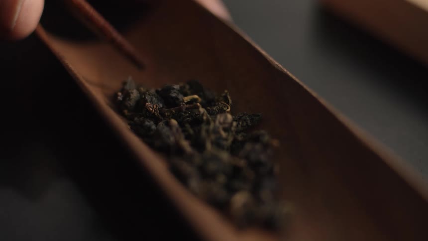 A person uses small wooden tweezers to pick up and arrange dried tea leaves in a narrow wooden tray on a dark surface in a close view