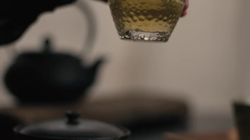 A close view shows a hand pouring golden tea from a dark round vessel into a clear textured glass pitcher on a wooden tray