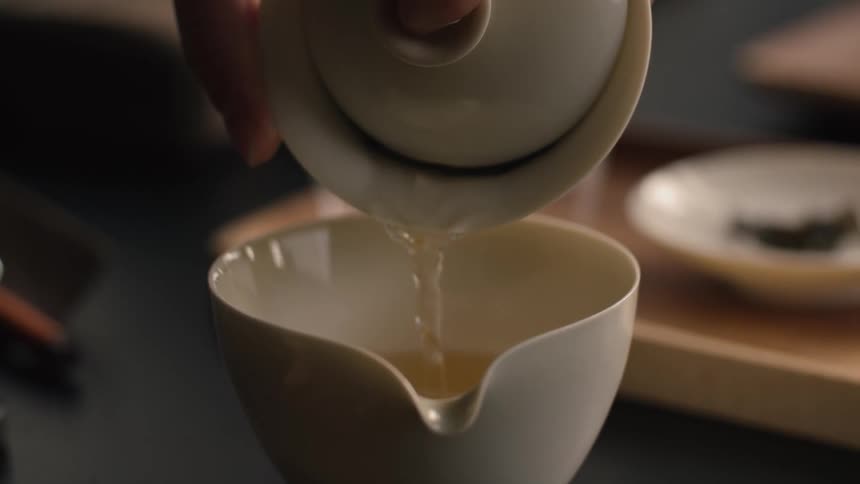 Hand pours light tea from white teapot into cup on wooden tray