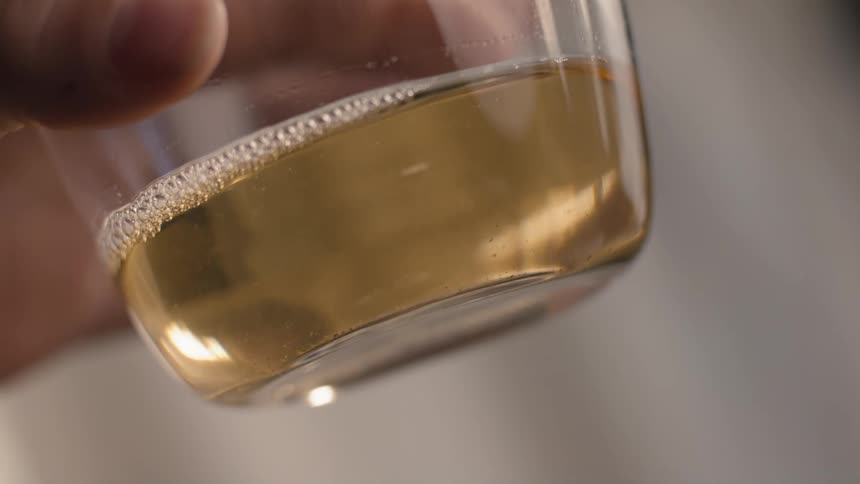 A person holds a clear glass containing a goldenamber colored liquid with a thin layer of foam at the surface gently swirling the drink in a tight closeup shot