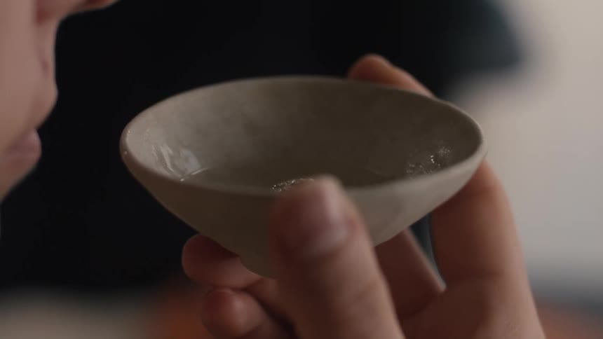 A close view shows a person bringing a small round beige ceramic cup of clear liquid up to their lips to drink