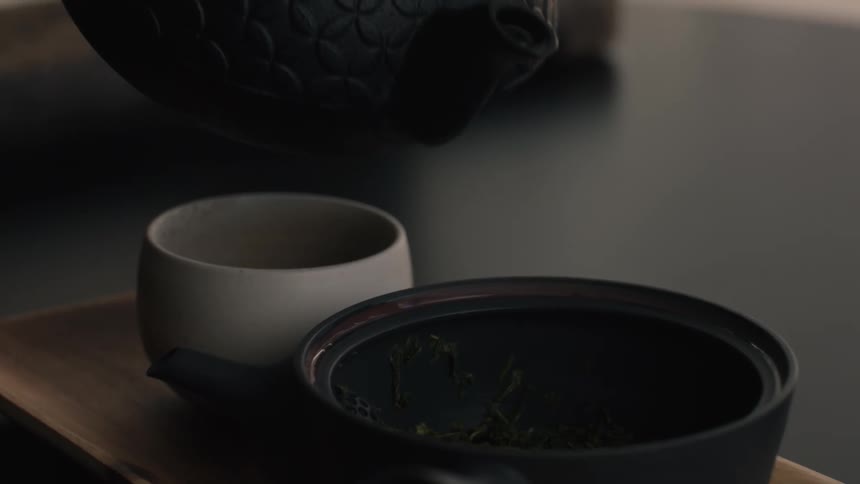 Hot water pours from black teapot over tea leaves in dark vessel on tray