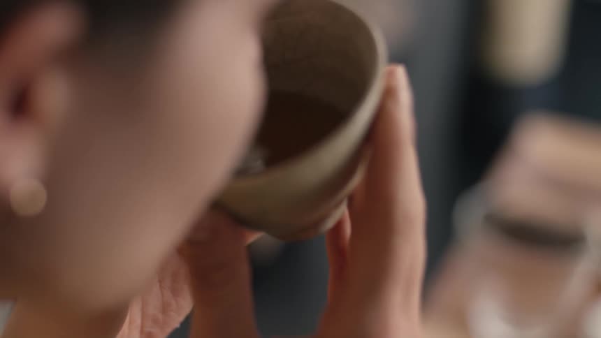 A close view shows a woman holding a small light colored ceramic cup with a cracked glaze containing brown liquid