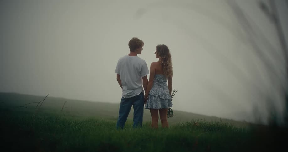 Two people holding hands face each other on grassy hill wrapped in soft mist and romantic ambiance