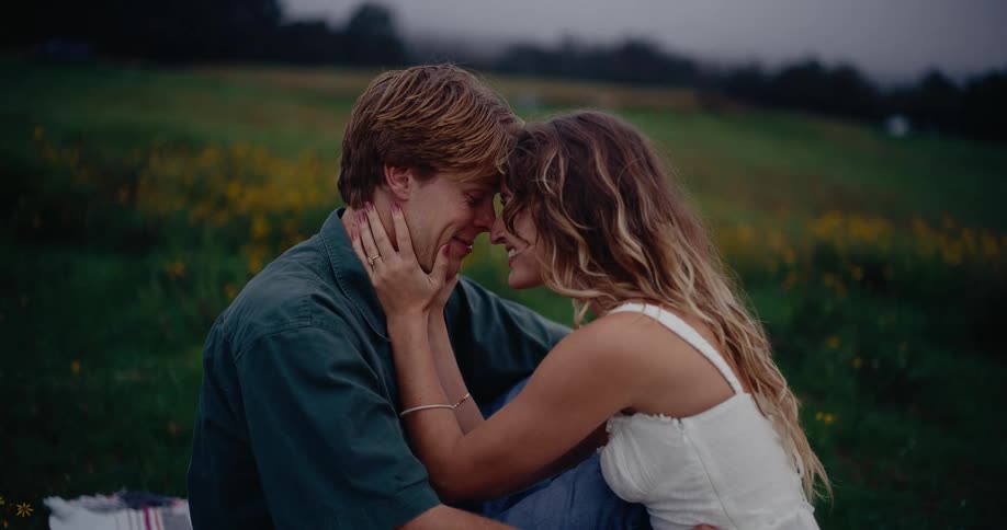 A couple sits close together in a foggy meadow filled with yellow flowers at dusk gazing at each other with affection their heads and hands touching in an intimate moment of connection