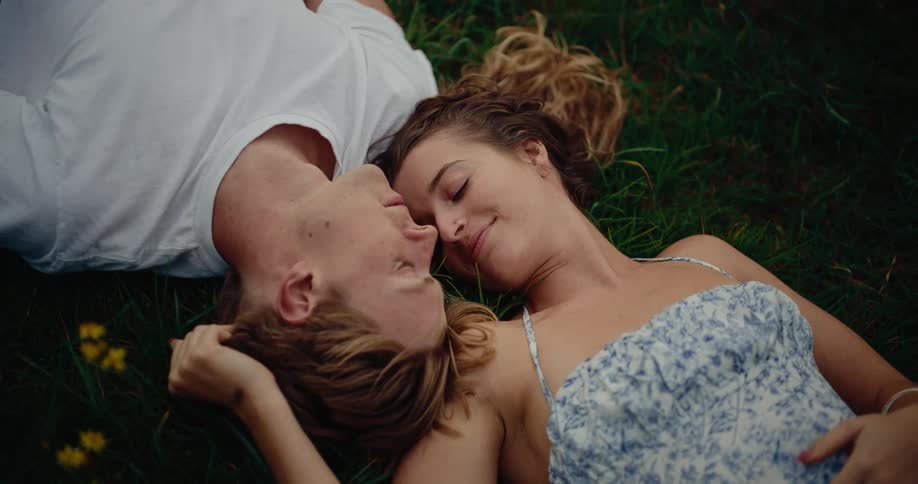 A young couple lies on a grass lawn at different angles their heads side by side as the woman gently rubs her hand on the back of the mans head