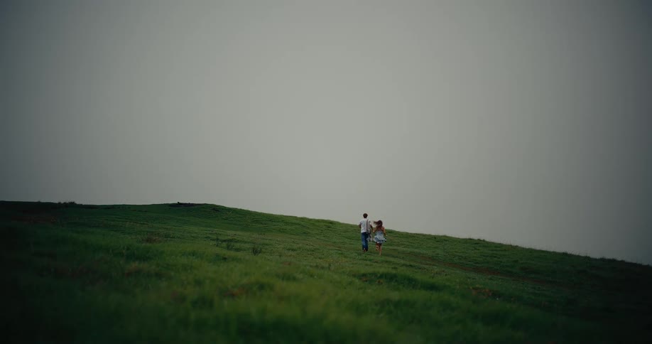 A man in a white shirt and a woman in a short dress walk hand in hand up a lush green hill under an overcast misty sky