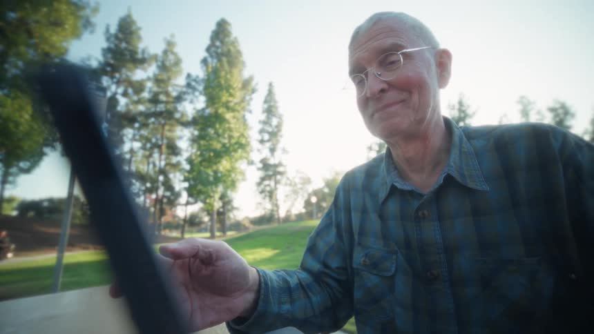 Elderly man smiles while using a device in the park during the daytime