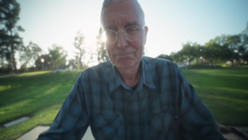 An elderly man wearing glasses sits on a bench in a public park at midday looking at his computer screen