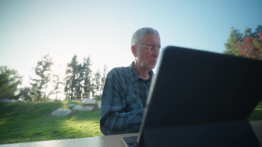 Man sits outside at a table with a laptop computer as he scrolls on his phone