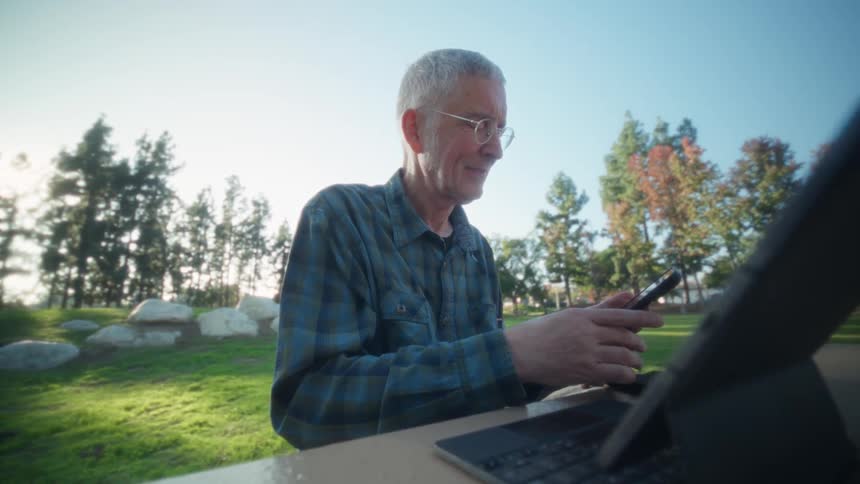 Elderly man wearing glasses and a plaid shirt using a smartphone sitting on a table in front of a laptop computer at a park outdoors during day time