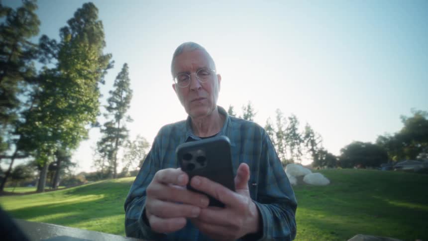 Man sits outside at a table with a laptop computer as he scrolls on his phone