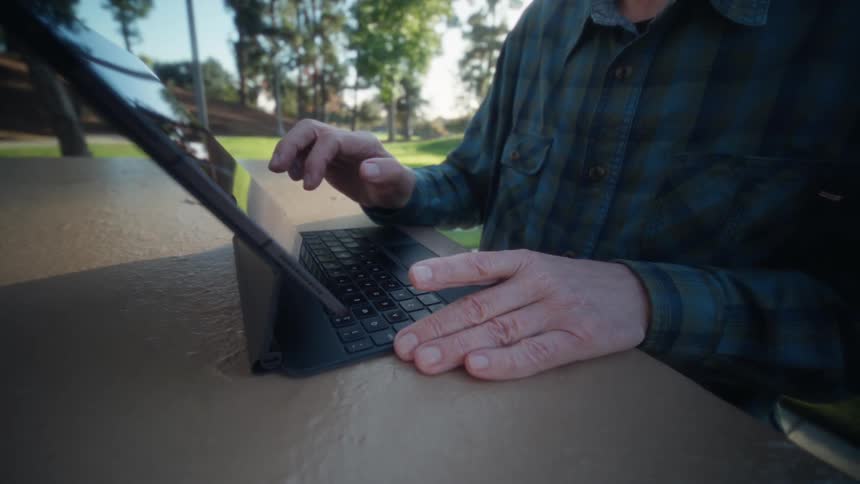 Person in blue plaid shirt interacts with tablet on outdoor surface surrounded by grass and trees