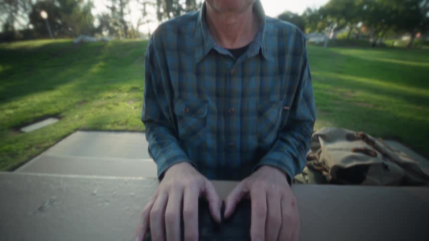 Person in blue plaid shirt types on tablet with keyboard on outdoor table beside beige bag