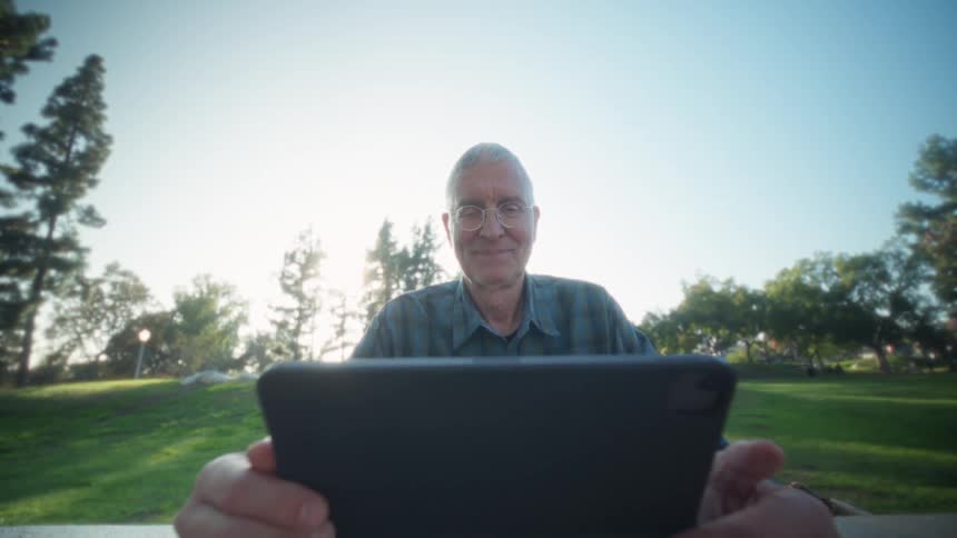 Older adult using tablet with both hands in treelined park under clear sky