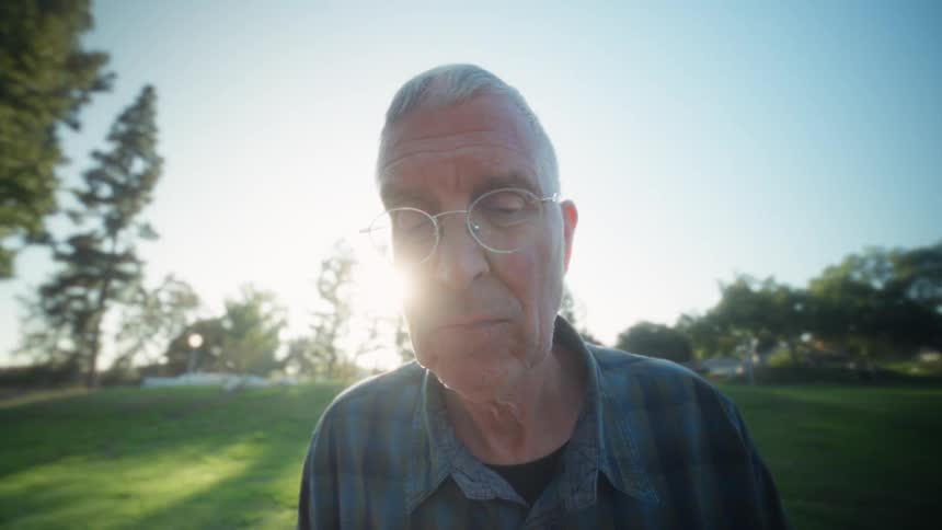 An older man with glasses wearing a plaid shirt intently working on a tablet at an outdoor park table on a sunny day before briefly looking up and then returning to his work
