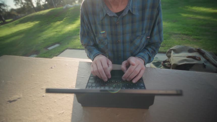 Person in blue plaid shirt types on tablet with keyboard on outdoor concrete table