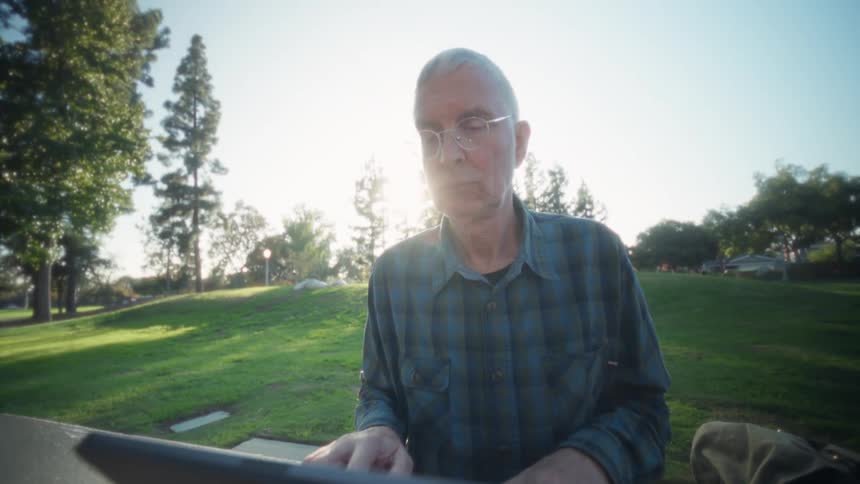 Elderly man wearing glasses and a plaid shirt using a tablet computer on a table at a park outdors during dusk or dawn