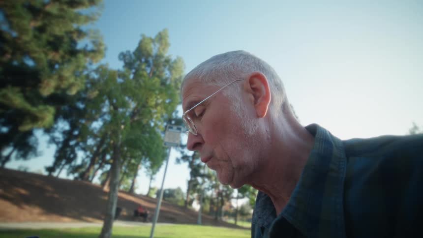 An older man wearing glasses and a plaid shirt works on a tablet attached to a keyboard sitting at a table in a sunny park with trees and grass visible in the background
