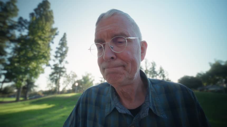 An older man wearing glasses and a plaid shirt works intently on a tablet computer outdoors at a park table