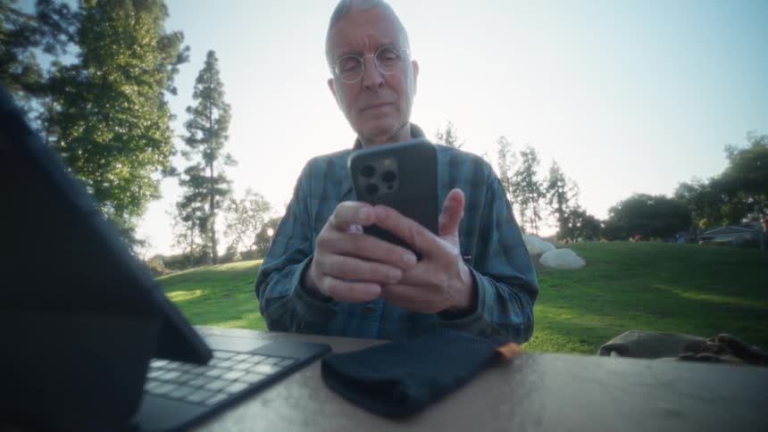 Elderly man sitting down at a park using a smartphone with a tablet computer in a wooden table in front of him outdoors during dusk or dawn