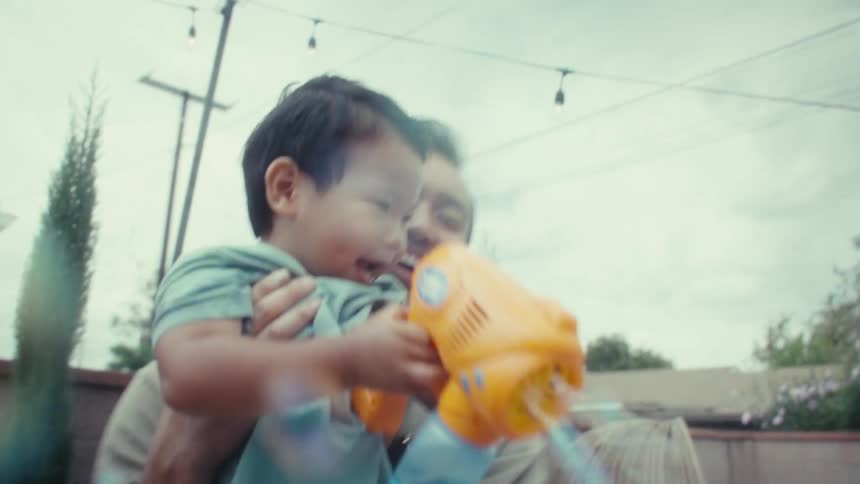 The man is holding the little boy who is shooting his bubble gun while the mother walks in the background