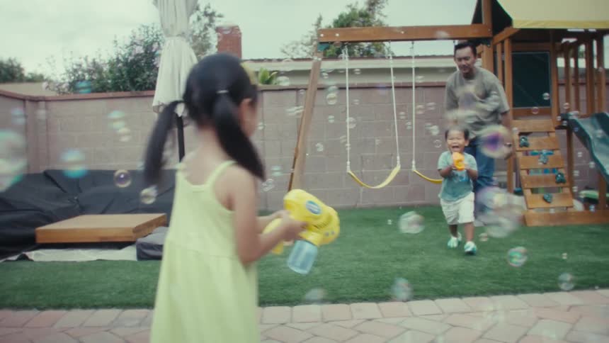 The boy and girl are playing with bubble guns inside of their back yard with their dad standing and watch and mom squatting doing something to the grass
