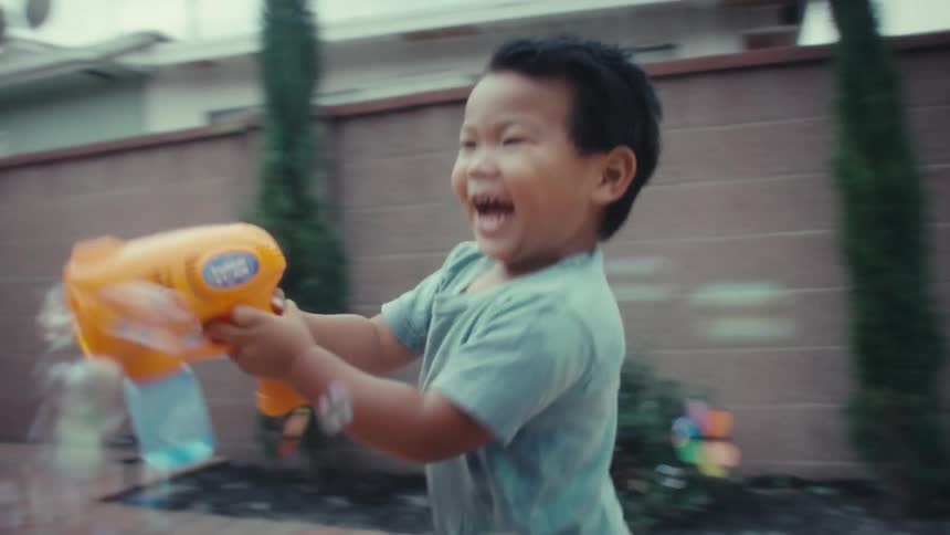 An excited boy in a light blue shirt runs on a paved backyard patio while shooting bubbles from an orange and yellow toy gun on a bright day