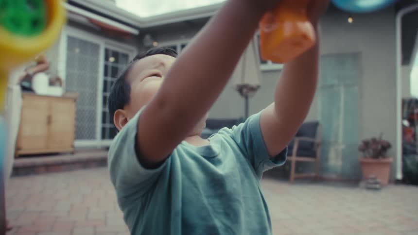 A smiling little boy in a teal shirt holding an orange and yellow toy bubble gun looking intently at the soap bubbles being produced in a backyard patio area