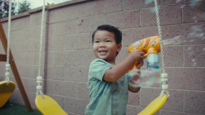 Small boy playing around with a bubble toy gun and running towards a female adult on a playground outdoors during day time