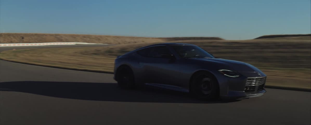 A sleek dark gray sports coupe drives quickly around a curve on an asphalt track under a clear blue sky casting a dark shadow on the road