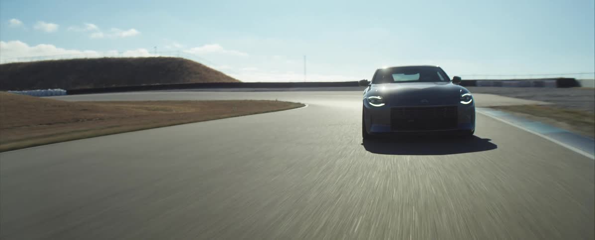 A dark gray sports car with its headlights on speeds down an asphalt race track on a bright sunny day with dry grass fields surrounding the course