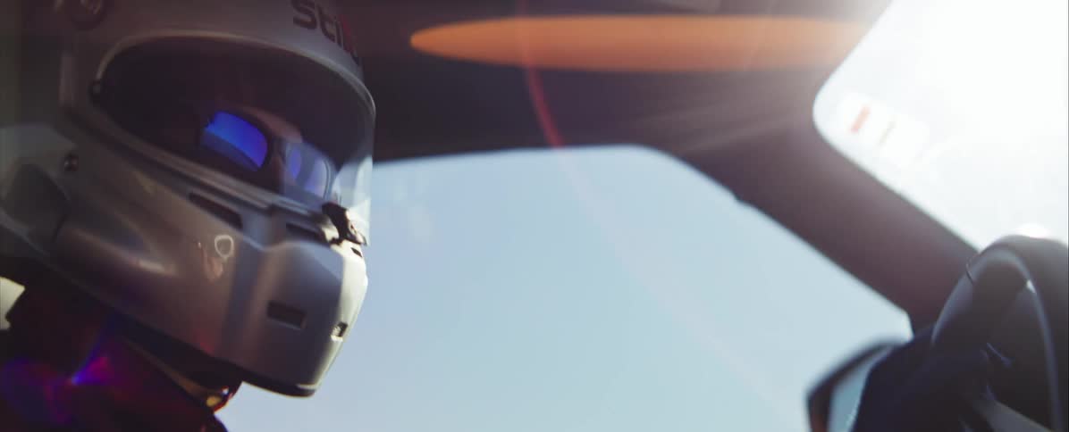 A close view shows a race car driver wearing a white helmet with a dark visor and red accents sitting in a cockpit with the sun flaring from the top right