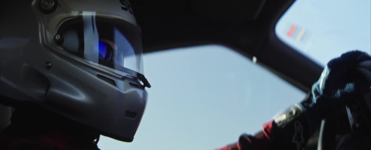 A closeup view from inside a car of a driver wearing a silver racing helmet looking forward with intense focus suggesting a highspeed motorsport event under a bright sky