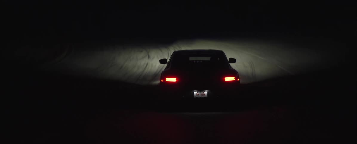 Tail lights of a black sports car glow in the darkness as it travels on a snow covered road at night