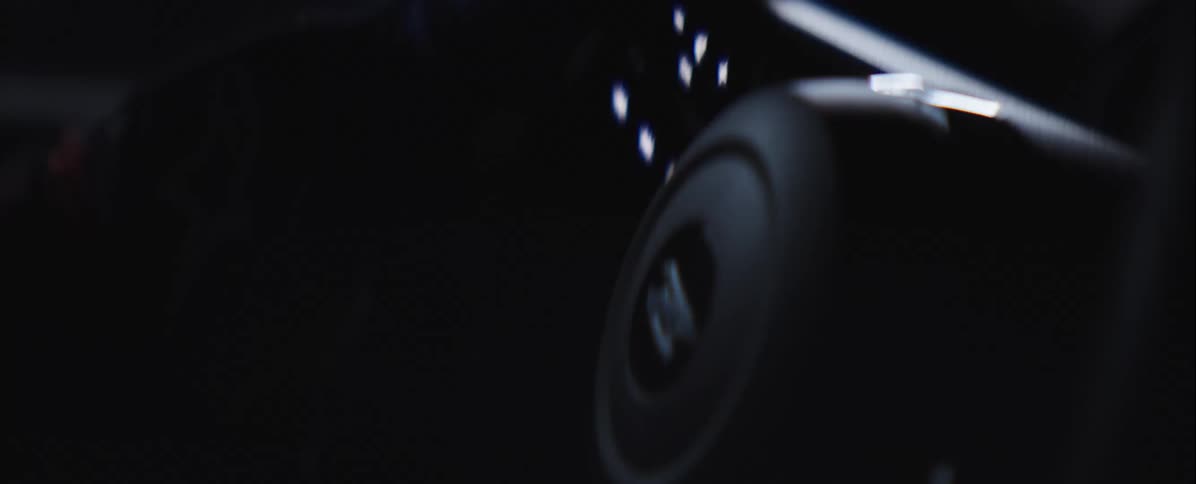 A tight lowlight shot focuses on the glowing circular control knob on a vehicles dashboard with small white indicator lights and hands with gloves driving on a dark environment