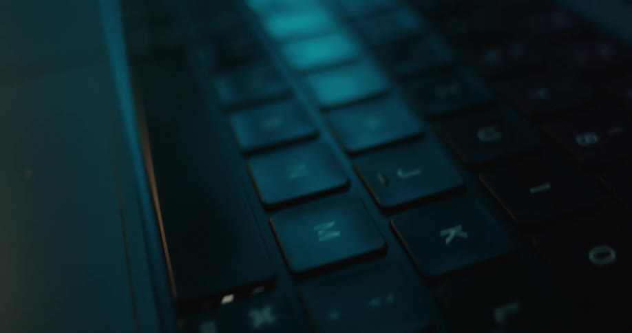 An extreme closeup of a laptop keyboard under dark moody lighting and a blue glow showing the black keys and white lettering in detail
