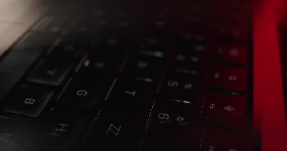 A close view of a dark computer keyboard with red ambient light reflecting off the shiny keys revealing some letters and numbers