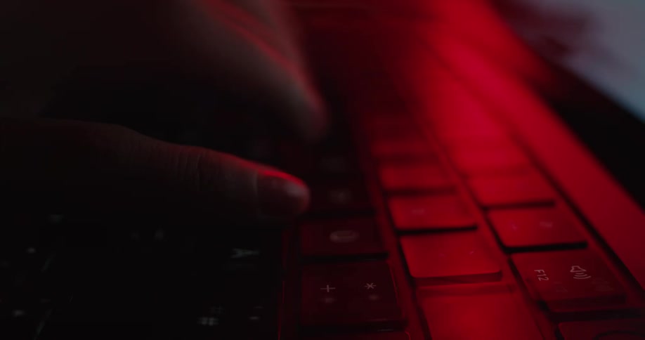 A closeup shot focuses on a persons fingers rapidly pressing keys on a keyboard which is dramatically illuminated by a deep dark red light