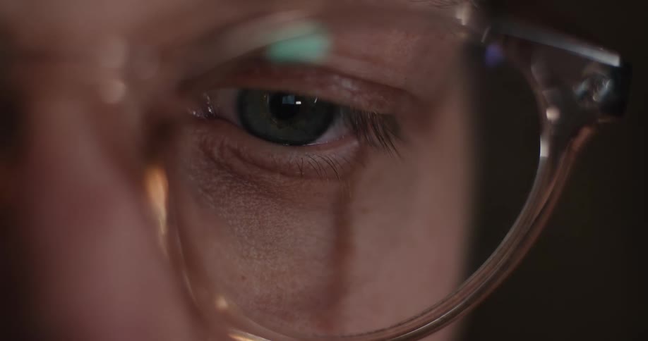 The man in the glasses is sitting in the dark looking at something that produces light as it is reflecting in his glasses