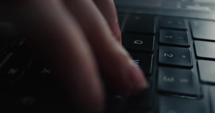 A close view shows a womans hands quickly typing on a black laptop keyboard under dim dramatic lighting