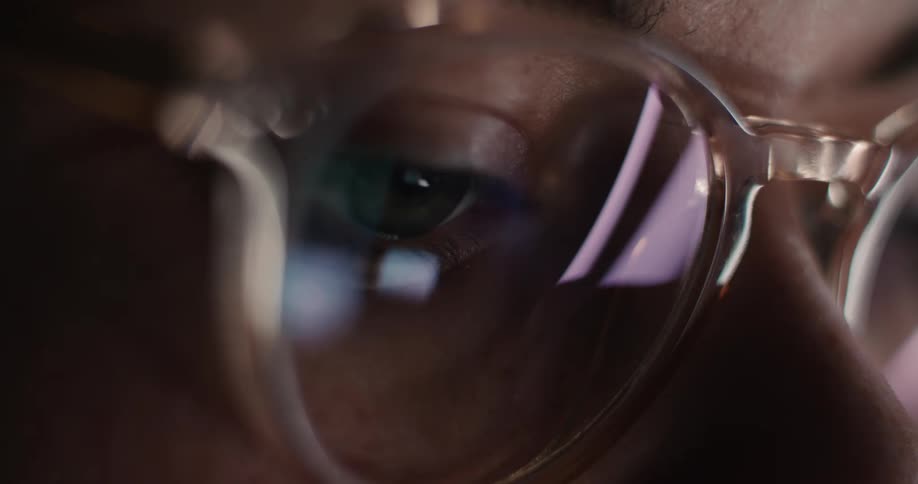 A very close view shows a womans eye and glasses reflecting the bright screen light of a laptop or tablet in a dark room