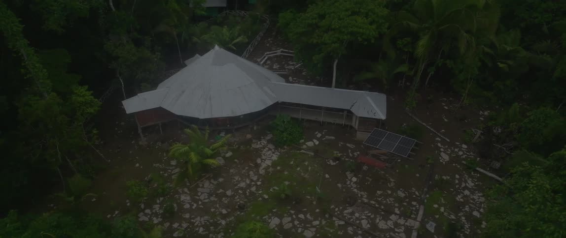An overhead slowzoom drone shot descends to reveal a large round metalroofed structure with attached buildings seemingly offgrid with solar panels situated on cleared rocky land surrounded by dense darkgreen tropical jungle