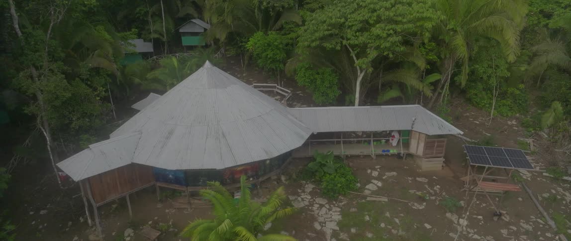 An aerial shot overlooks a circular structure with a large conical metal roof situated in a jungle clearing a solar panel is visible next to a covered porch area and the building is surrounded by dense tropical foliage