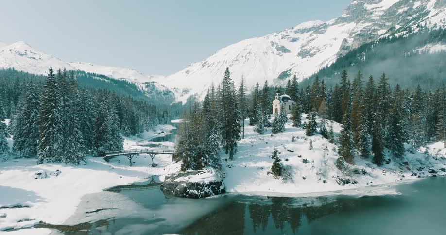 A stone building sits among snowy trees on an island in a partially frozen mountain lake