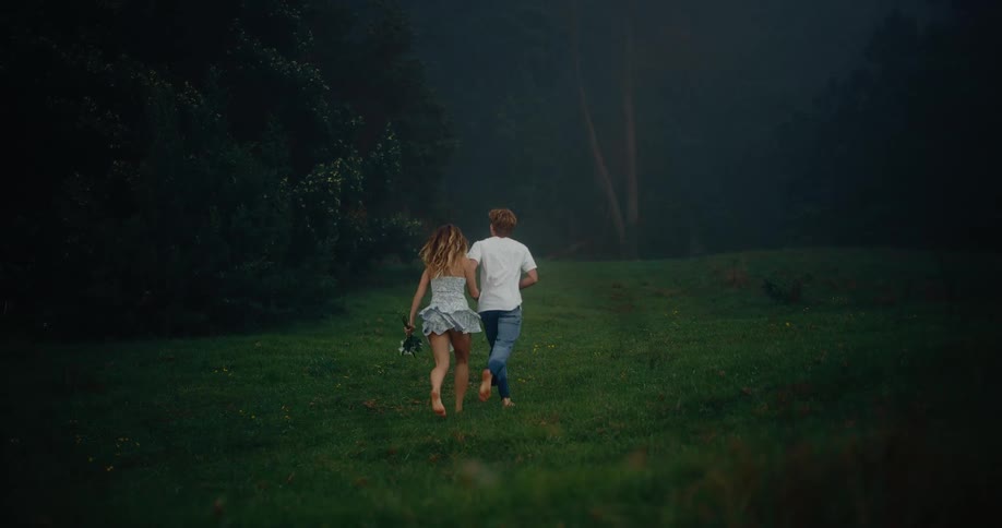 A couple the woman carrying flowers and wearing a light patterned dress and the man in a white shirt and blue jeans runs barefoot across a dark green grassy field towards the shadowy edge of a dense forest